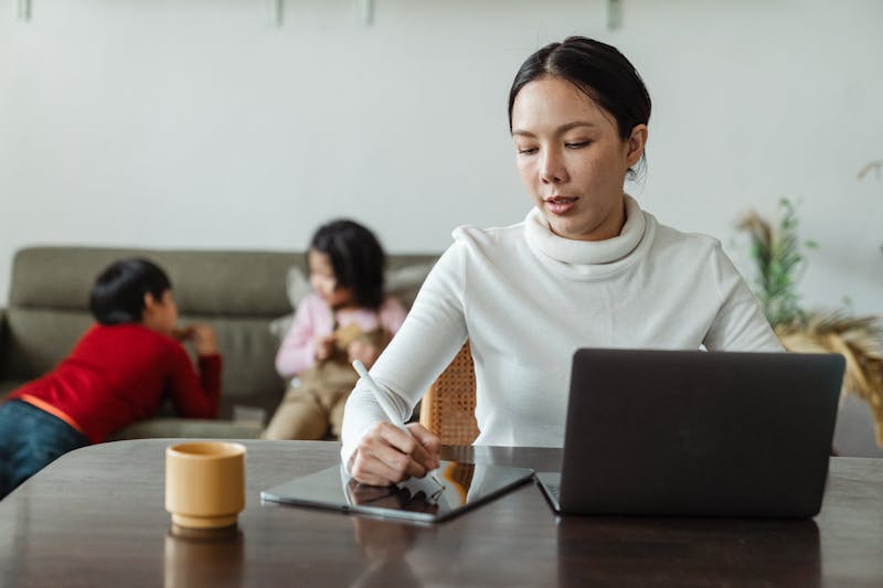 Working ethnic mother using tablet and laptop at home with playing kids on background